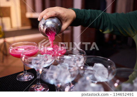 Mood setting with lively drink preparation at upscale bar, Elegant scene of bartender pouring colorful cocktail amidst festive warm lighting and guests in background 134652491