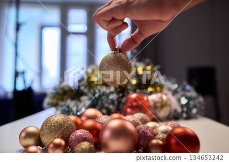 Hands arranging Christmas New Year baubles on white table with tinsel and warm bokeh lights, metallic red gold pink palette, careful styling by home decorator, festive craft mood 134653242