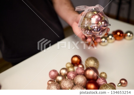 Hands arranging Christmas New Year baubles on white table with tinsel and warm bokeh lights, metallic red gold pink palette, careful styling by home decorator, festive craft mood 134653254