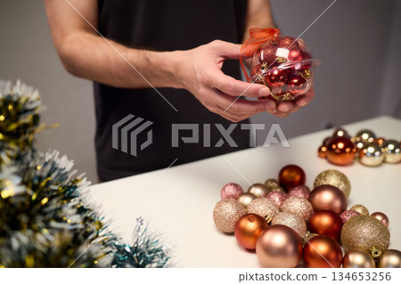 Hands arranging Christmas New Year baubles on white table with tinsel and warm bokeh lights, metallic red gold pink palette, careful styling by home decorator, festive craft mood 134653256