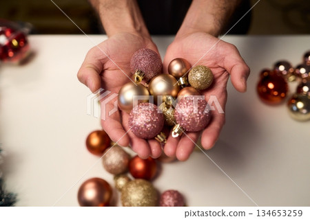 Hands arranging Christmas New Year baubles on white table with tinsel and warm bokeh lights, metallic red gold pink palette, careful styling by home decorator, festive craft mood 134653259