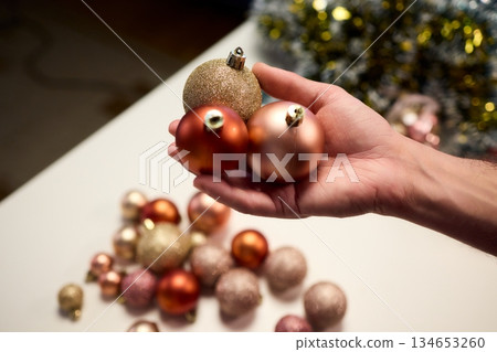 Hands arranging Christmas New Year baubles on white table with tinsel and warm bokeh lights, metallic red gold pink palette, careful styling by home decorator, festive craft mood 134653260