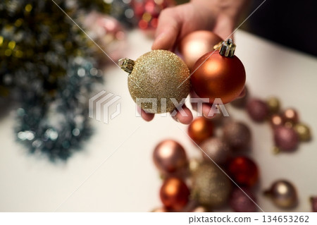 Hands arranging Christmas New Year baubles on white table with tinsel and warm bokeh lights, metallic red gold pink palette, careful styling by home decorator, festive craft mood 134653262