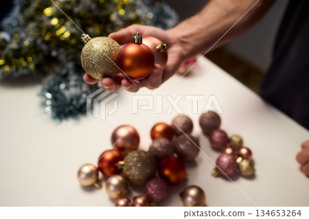 Hands arranging Christmas New Year baubles on white table with tinsel and warm bokeh lights, metallic red gold pink palette, careful styling by home decorator, festive craft mood Hands arranging Christmas New Year baubles on white table with tinsel and warm bokeh lights, metallic red gold pink palette, careful styling by home decorator, festive craft mood 134653264