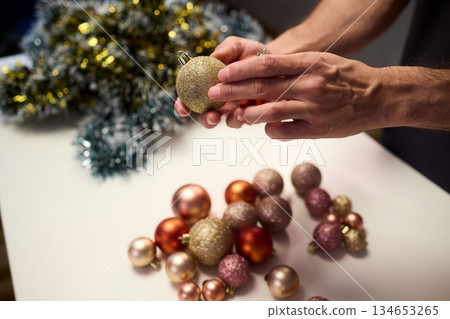 Hands arranging Christmas New Year baubles on white table with tinsel and warm bokeh lights, metallic red gold pink palette, careful styling by home decorator, festive craft mood Hands arranging Christmas New Year baubles on white table with tinsel and warm bokeh lights, metallic red gold pink palette, careful styling by home decorator, festive craft mood 134653265