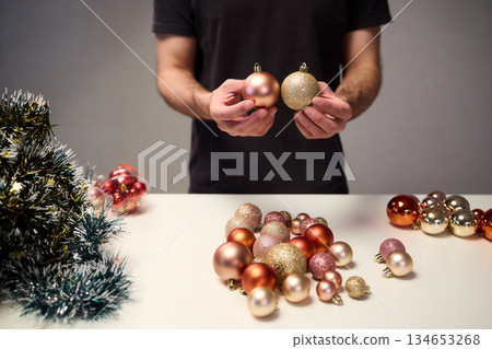 Hands arranging Christmas New Year baubles on white table with tinsel and warm bokeh lights, metallic red gold pink palette, careful styling by home decorator, festive craft mood 134653268