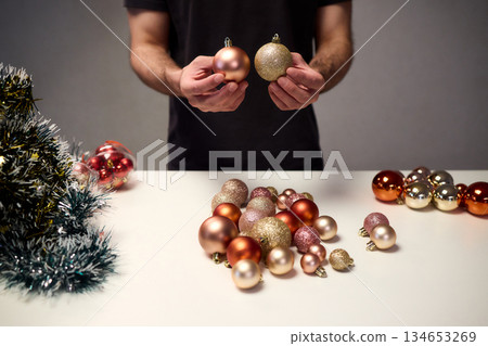 Hands arranging Christmas New Year baubles on white table with tinsel and warm bokeh lights, metallic red gold pink palette, careful styling by home decorator, festive craft mood 134653269