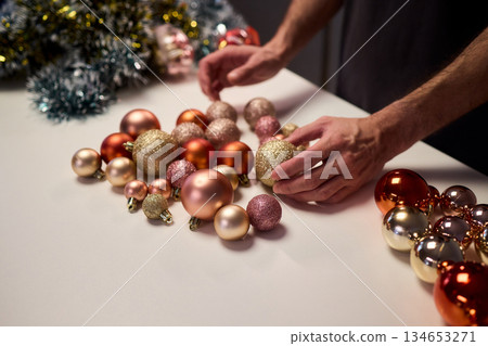 Hands arranging Christmas New Year baubles on white table with tinsel and warm bokeh lights, metallic red gold pink palette, careful styling by home decorator, festive craft mood 134653271