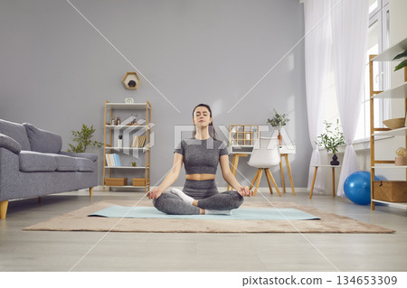 Young yogi calm sporty attractive woman practicing yoga at home doing Ardha Padmasana exercise 134653309