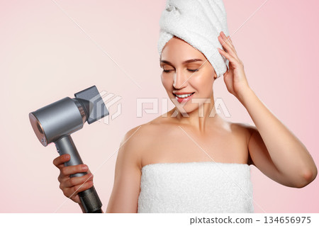 Woman in towel holds hair dryer in front of pink background with bright lighting 134656975