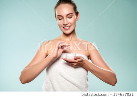 Woman applying cream while holding a jar in a light setting with a simple background Woman applying cream while holding a jar in a light setting with a simple background 134657021