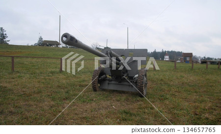 field howitzer positioned on open grass with long barrel and carriage visible, countryside houses on horizon give context to decommissioned training ground turned memorial display under muted sky. 134657765