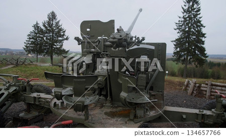 decommissioned antiaircraft gun with exposed controls and weathered paint, stationed on grassy hill beside pine trees, visible seats, gears and cables create somber industrial museum atmosphere 134657766