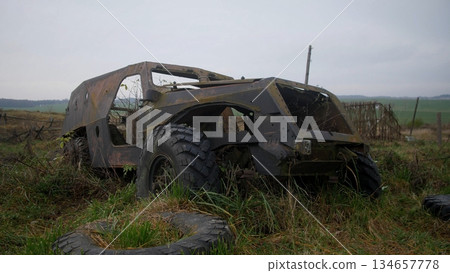 burned armored vehicle stranded in overgrown field, scorched hull and exposed interior with flat tire, charred metal and scattered debris convey post-conflict decay and eerie desolation under cloudy 134657778