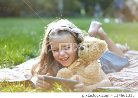 Small kid girl looking in her mobile phone together with her favorite teddy bear toy outdoors in summer park. Small kid girl looking in her mobile phone together with her favorite teddy bear toy outdoors in summer park. 134661333