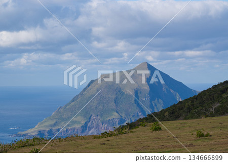 從八丈島歡樂牧場可以欣賞到懸崖、山脈和大海的景色。藍天白雲籠罩著整個島嶼和八丈小島。 從八丈島歡樂牧場可以欣賞到懸崖、山脈和大海的景色。藍天白雲籠罩著整個島嶼和八丈小島。 134666899