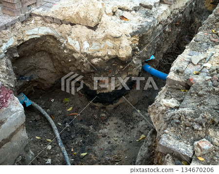 Deep excavation trench revealing blue corrugated pipe and grey utility line, signifying ongoing urban infrastructure construction, essential repair, and groundwork Deep excavation trench revealing blue corrugated pipe and grey utility line, signifying ongoing urban infrastructure construction, essential repair, and groundwork 134670206