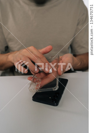 Vertical cropped shot of man wiping transparent silicone phone case with small white cloth over black smartphone, close-up domestic routine emphasizing hygiene, protection and device care. 134670751