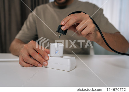 Man holding power plug, connecting it to smart socket inserted in power strip, demonstrating modern smarthome technology for energy management and automation, close-up. Man holding power plug, connecting it to smart socket inserted in power strip, demonstrating modern smarthome technology for energy management and automation, close-up. 134670752