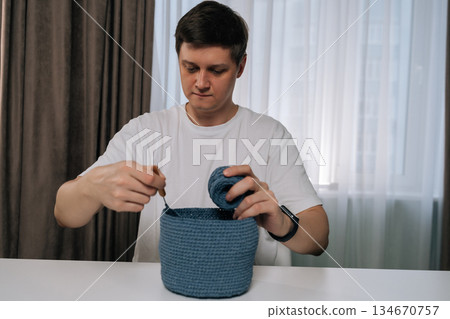 Portrait of young man focused on crocheting blue yarn basket at home, practicing traditional needlework and enjoying mindful, creative hobby that blends skill, relaxation, handmade design. Portrait of young man focused on crocheting blue yarn basket at home, practicing traditional needlework and enjoying mindful, creative hobby that blends skill, relaxation, handmade design. 134670757