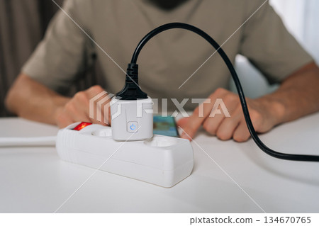 Cropped shot of person wirelessly setting up smart socket device plugged into power strip, interacting with smartphone display for home automation and remote control of appliances. 134670765