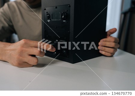 Close-up cropped shot of unrecognizable musician male connecting power cable to black monitor speaker, setting up audio equipment on white desk, preparing home studio or entertainment system for use. 134670770