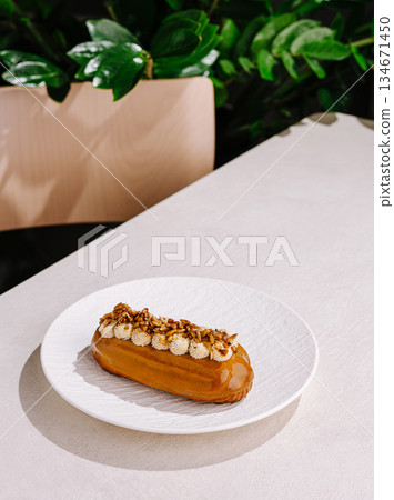 Sweet caramel eclair with nuts on a white plate in a cafe setting Sweet caramel eclair with nuts on a white plate in a cafe setting 134671450