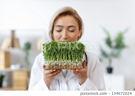 A woman holds a tray of fresh microgreens, likely pea shoots, close to her face, possibly smelling them. 134672024