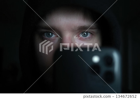 Individual with blue eyes stares directly at the camera while wearing a hood in a dark setting. The focus is on the face and eyes Individual with blue eyes stares directly at the camera while wearing a hood in a dark setting. The focus is on the face and eyes 134672652
