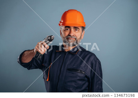 Repairer, holding drill, in hardhat. Senior man is standing in the studio against background Repairer, holding drill, in hardhat. Senior man is standing in the studio against background 134673198