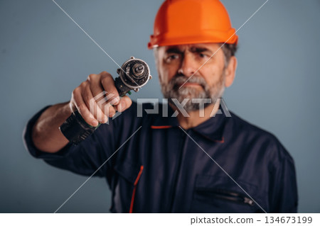 Repairer, holding drill, in hardhat. Senior man is standing in the studio against background Repairer, holding drill, in hardhat. Senior man is standing in the studio against background 134673199