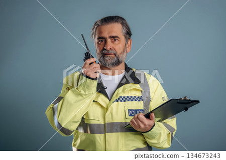 Talking into walkie talkie, police officer. Senior man is standing in the studio against background Talking into walkie talkie, police officer. Senior man is standing in the studio against background 134673243
