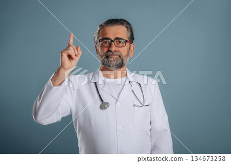 Professional doctor in coat. Senior man is standing in the studio against background Professional doctor in coat. Senior man is standing in the studio against background 134673258