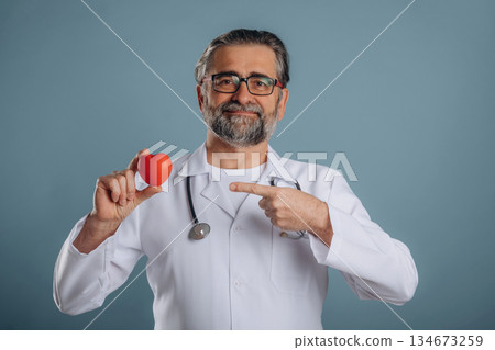 Stethoscope on neck, holding little heart. Senior man is standing in the studio against background 134673259