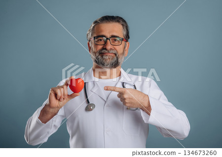 Stethoscope on neck, holding little heart. Senior man is standing in the studio against background 134673260