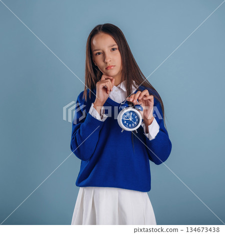 With blue clock in hand. Young schoolgirl in uniform is against background 134673438