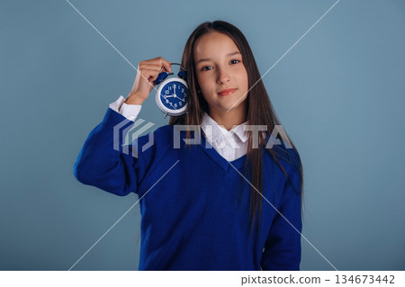 With blue clock in hand. Young schoolgirl in uniform is against background With blue clock in hand. Young schoolgirl in uniform is against background 134673442