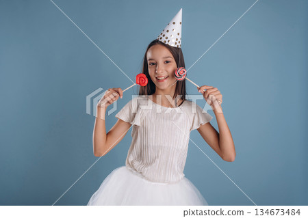 Birthday hat, with sweet candies. Young girl in white dress is against background 134673484