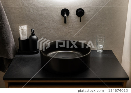 A modern bathroom sink with a round black basin on a dark countertop. There are black wall-mounted faucets, a glass, and a soap dispenser nearby. A modern bathroom sink with a round black basin on a dark countertop. There are black wall-mounted faucets, a glass, and a soap dispenser nearby. 134674013
