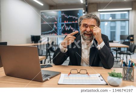 Expressive senior male office worker is with crypto coin in hand before the eye, smiling Expressive senior male office worker is with crypto coin in hand before the eye, smiling 134674197