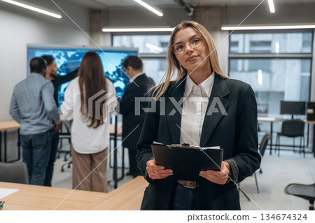 Woman in glasses is standing with notepad. Group of office workers are indoors together Woman in glasses is standing with notepad. Group of office workers are indoors together 134674324