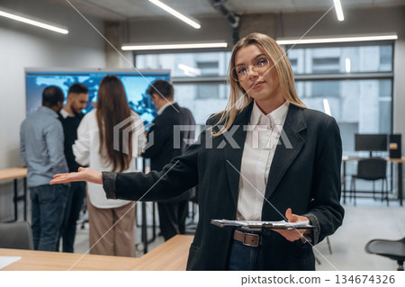 Woman in glasses is standing with notepad. Group of office workers are indoors together Woman in glasses is standing with notepad. Group of office workers are indoors together 134674326
