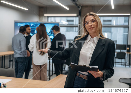 Woman in glasses is standing with notepad. Group of office workers are indoors together Woman in glasses is standing with notepad. Group of office workers are indoors together 134674327