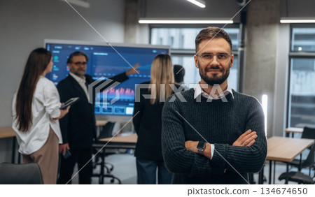 With arms crossed, standing against colleagues. Group of office workers are indoors together With arms crossed, standing against colleagues. Group of office workers are indoors together 134674650