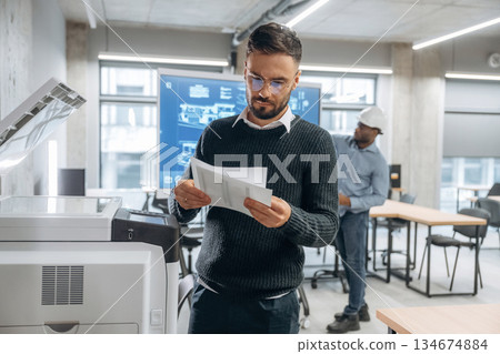 Two office workers are standing, man is with documents near printer Two office workers are standing, man is with documents near printer 134674884