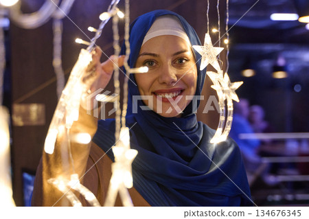 Woman wearing hijab enjoys decorations during Ramadan celebration in a festive setting with lights and stars Woman wearing hijab enjoys decorations during Ramadan celebration in a festive setting with lights and stars 134676345