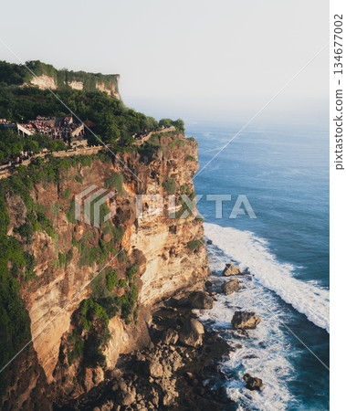 Uluwatu cliff temple Bali rising above Indian Ocean, dramatic limestone coastline with tourists at viewpoint, sacred landmark, tropical travel destination, ocean waves, spiritual atmosphere at sunset 134677002