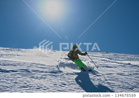 Skier skiing off piste through fresh powder on a sunny mountain slope, kicking up snow as she descends, creating a dynamic and energetic winter sports action scene under a clear blue sky 134678589