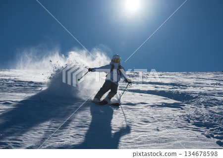 Skier skiing off piste through fresh powder on a sunny mountain slope, kicking up snow as she descends, creating a dynamic and energetic winter sports action scene under a clear blue sky Skier skiing off piste through fresh powder on a sunny mountain slope, kicking up snow as she descends, creating a dynamic and energetic winter sports action scene under a clear blue sky 134678598