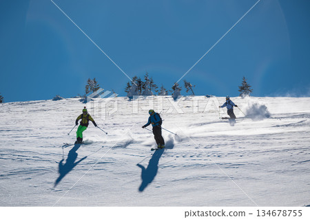 Three skiers carve down a sunny, powder-covered mountain slope, kicking up fresh snow under a clear blue sky, creating a dynamic winter sports action scene Three skiers carve down a sunny, powder-covered mountain slope, kicking up fresh snow under a clear blue sky, creating a dynamic winter sports action scene 134678755